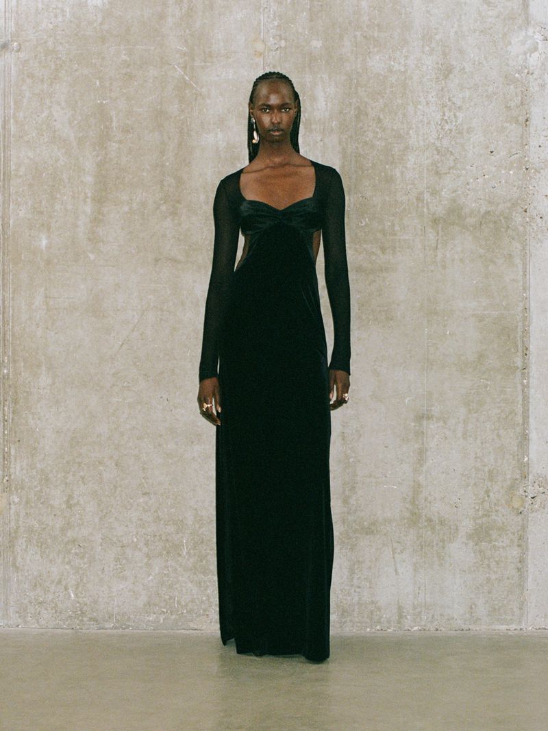 Elegant full-length studio shot of a Black woman in a long black evening gown with mesh sleeves and detail, against a textured grey wall.