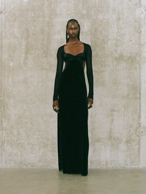 Elegant full-length studio shot of a Black woman in a long black evening gown with mesh sleeves and detail, against a textured grey wall.