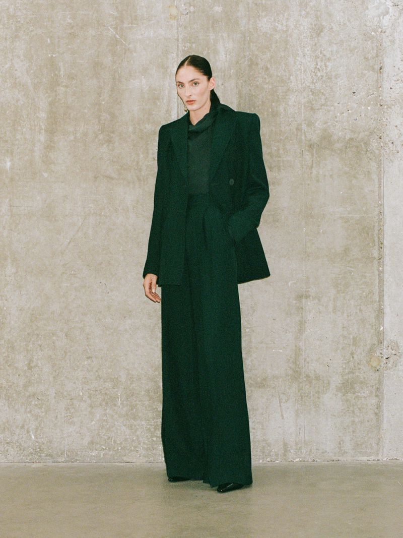 Indoor fashion shot of a stylish woman in an emerald green blazer and wide-leg pants, posing against a textured neutral wall.