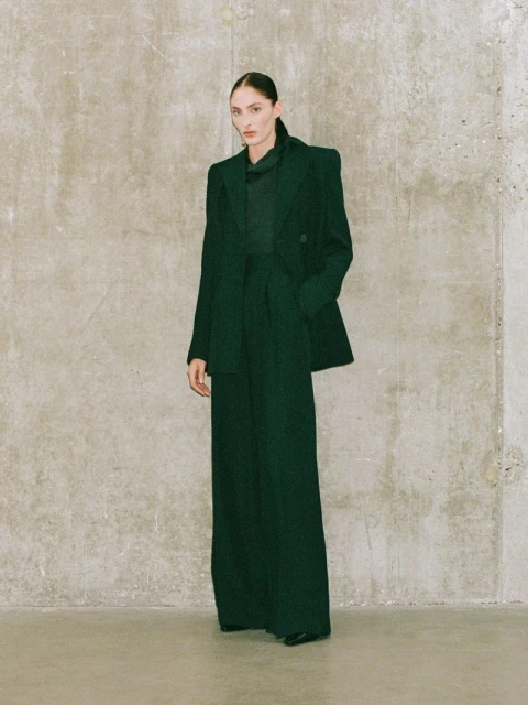 Indoor fashion shot of a stylish woman in an emerald green blazer and wide-leg pants, posing against a textured neutral wall.