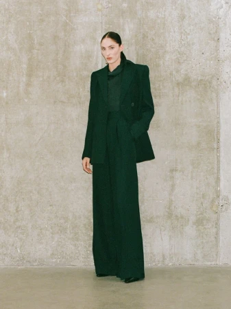 Indoor fashion shot of a stylish woman in an emerald green blazer and wide-leg pants, posing against a textured neutral wall.