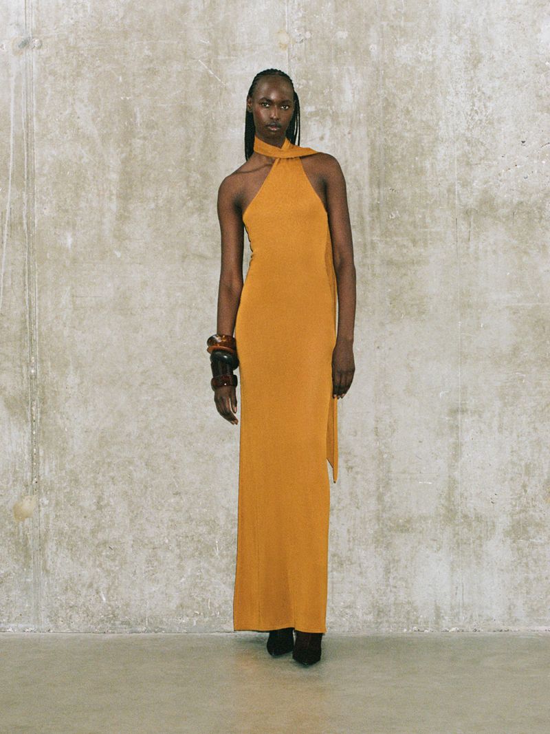 Elegant woman in a golden halter gown posing against a textured concrete wall, accessorized with chunky brown bracelets, fashion portrait.