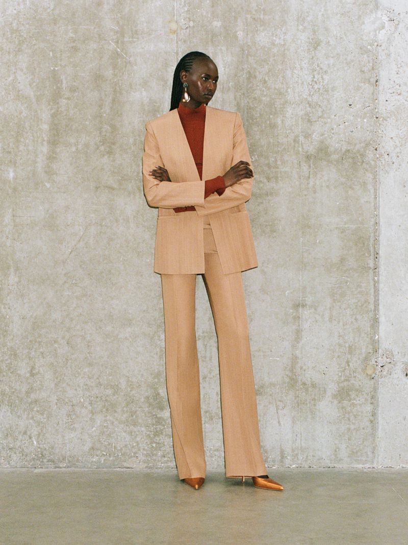 Fashion portrait of an African-descent adult woman wearing a beige pantsuit with a red turtleneck, posing in a photo studio.