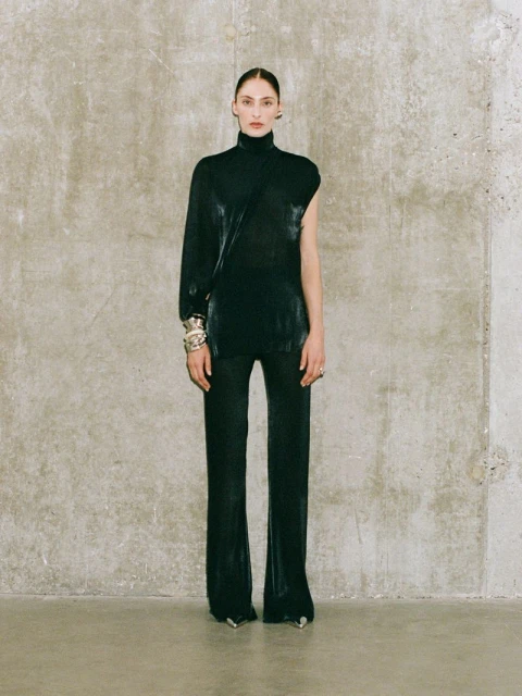 Woman wearing a black, one-sleeved top and matching pants poses in front of a concrete wall. She is adorned with silver bracelets.