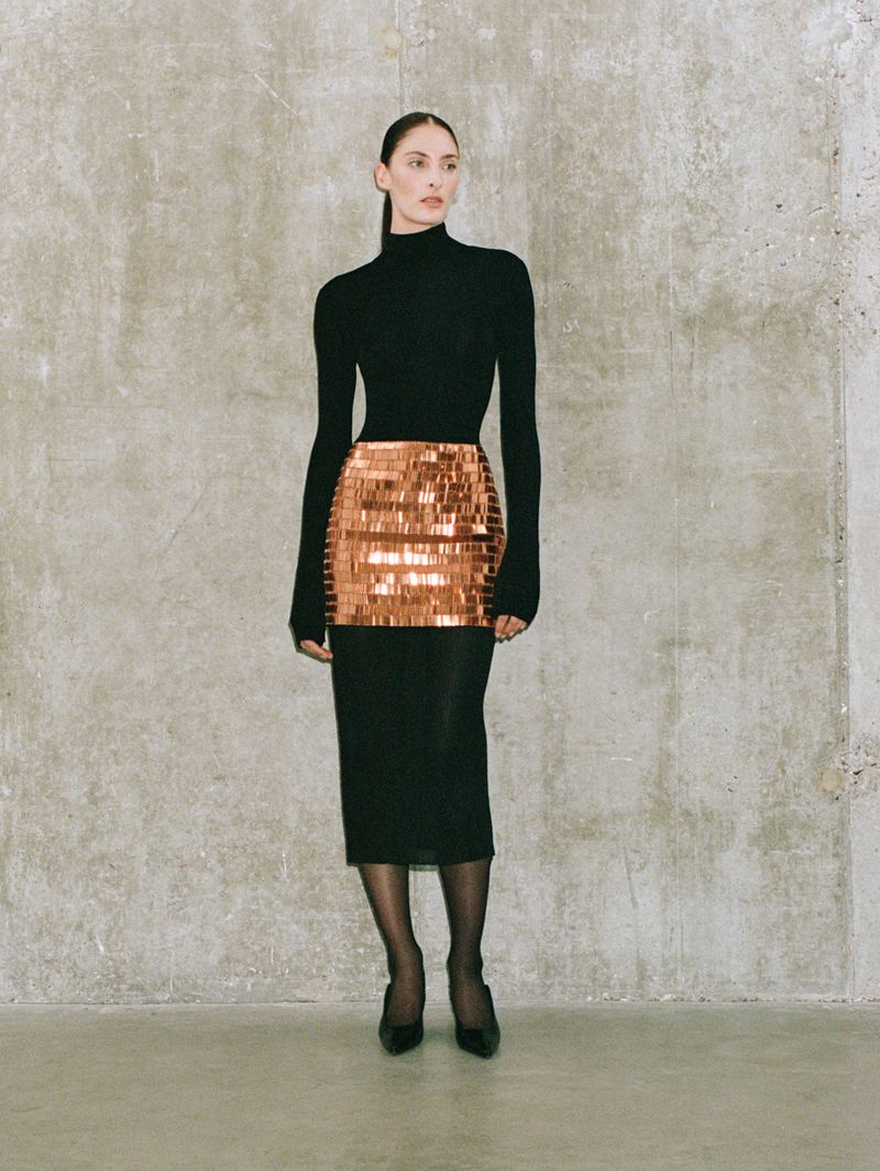 Woman in a black turtleneck and sequined mini skirt poses against a textured grey wall. The shot is full length and in a studio setting.