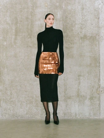 Woman in a black turtleneck and sequined mini skirt poses against a textured grey wall. The shot is full length and in a studio setting.
