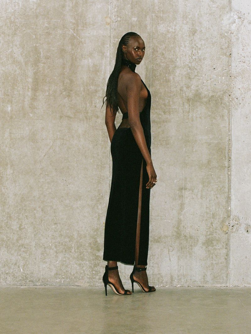 A slender Black woman posing in a black dress and heels against a textured concrete wall in a fashion forward presentation.