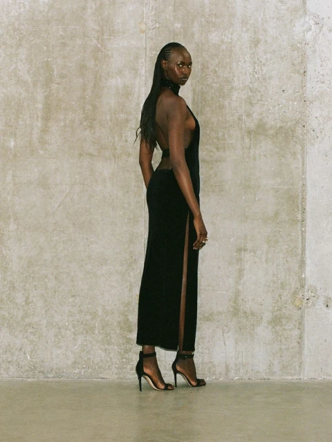 A slender Black woman posing in a black dress and heels against a textured concrete wall in a fashion forward presentation.