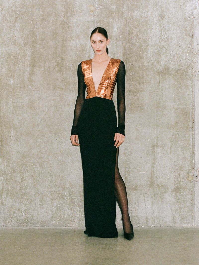 Woman poses in a black gown with a metallic bodice and long sleeves against a neutral wall for a fashion editorial photoshoot.