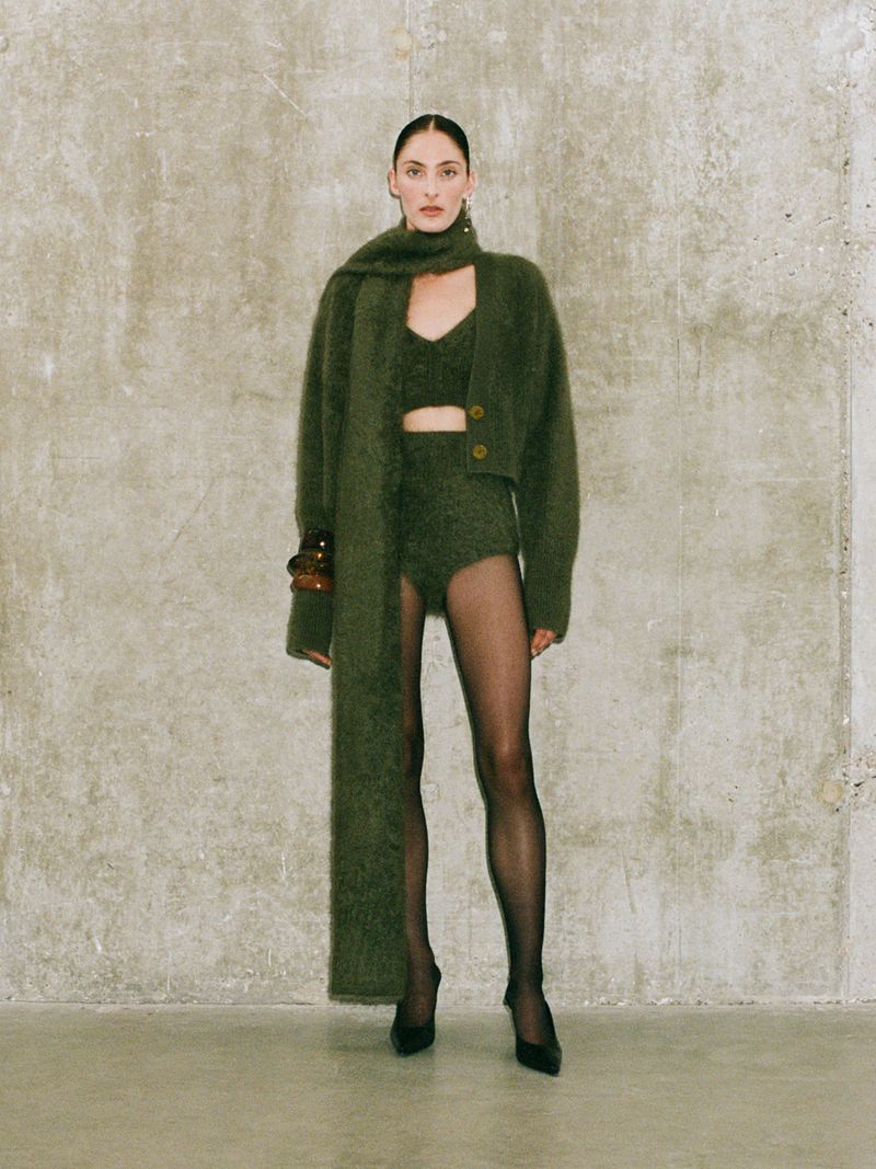 Elegant woman in a fuzzy, olive-green outfit posing in front of a textured grey wall in a fashion editorial, holding a unique handbag.