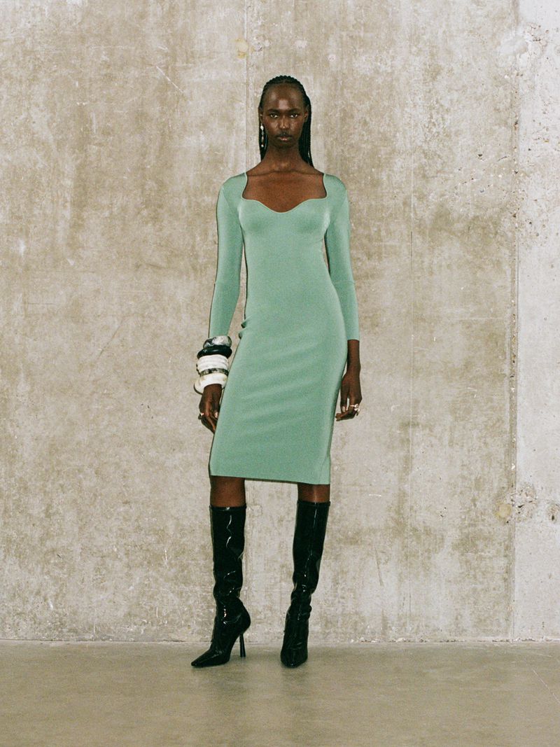 A black female model in a green dress and black patent leather boots stands against a textured neutral wall in a studio setting.
