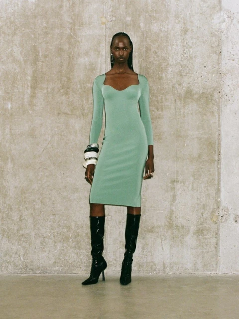 A black female model in a green dress and black patent leather boots stands against a textured neutral wall in a studio setting.