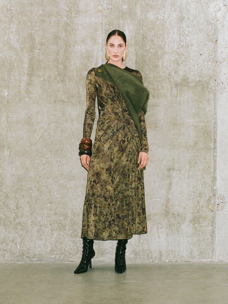 Fashion shot of a woman in a long-sleeved top, brown pants, and a long green scarf, posing against a concrete wall, looking at the camera.