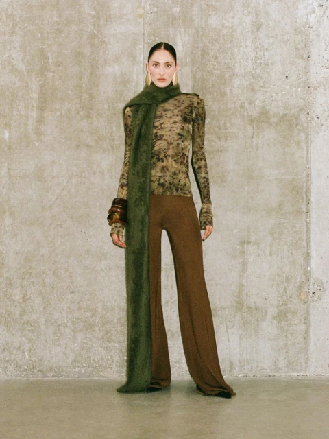 Fashion shot of a woman in a long-sleeved top, brown pants, and a long green scarf, posing against a concrete wall, looking at the camera.