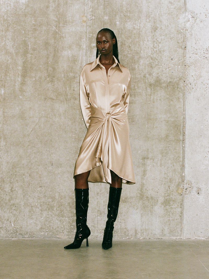 Full-length shot of a Black woman in a tied beige shirt-dress with black patent leather boots against a textured grey wall.