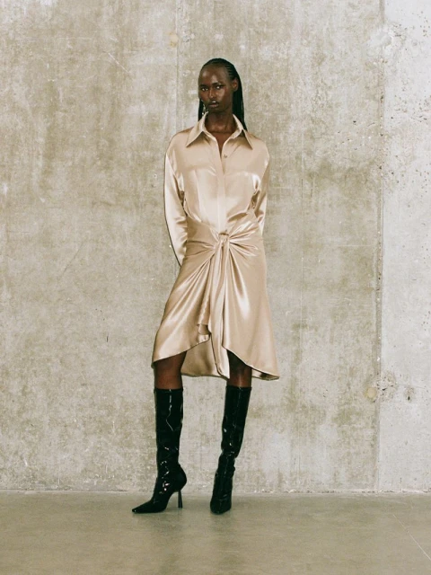 Full-length shot of a Black woman in a tied beige shirt-dress with black patent leather boots against a textured grey wall.