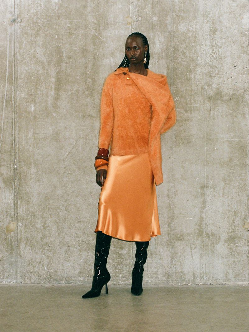 Woman in orange sweater and satin skirt poses against mottled grey wall indoors wearing knee high boots and ornate wrist cuff.