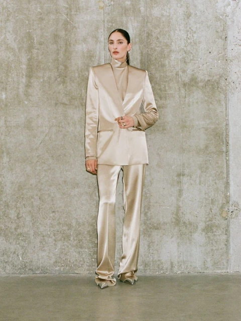 Woman poses in a stylish champagne-colored suit against a textured grey wall, showcasing modern, elegant fashion in a studio setting.