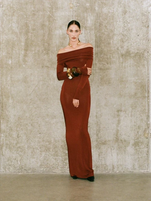 A woman in a long sleeve, off-the-shoulder brick red dress stands against a mottled gray wall, accessorized with a gold necklace and bracelets.