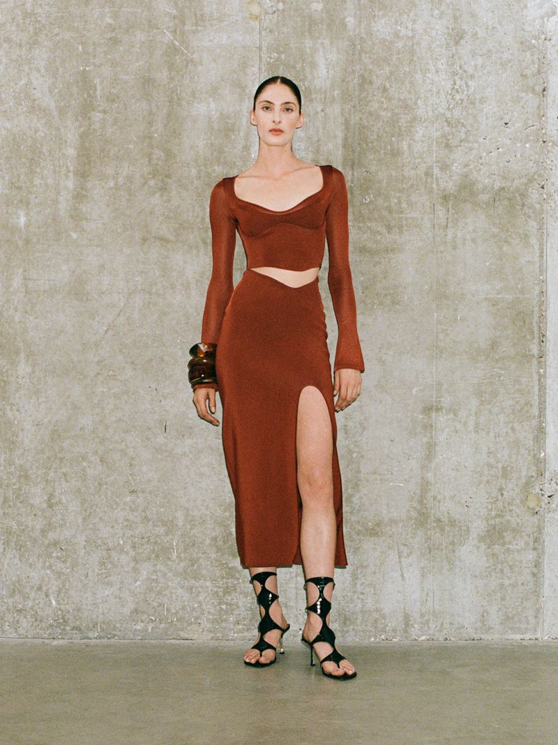 A poised woman showcases a rust-colored knit crop top and matching split midi skirt against a neutral backdrop, complemented by bold bracelets and black sandals.
