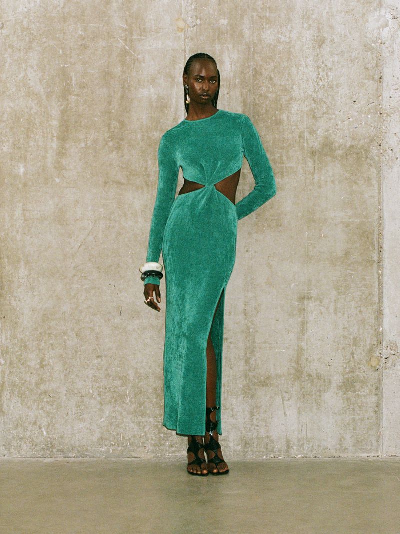 Adult woman posing in a full-length teal dress with cut-outs against a concrete backdrop in a fashion editorial style photo.