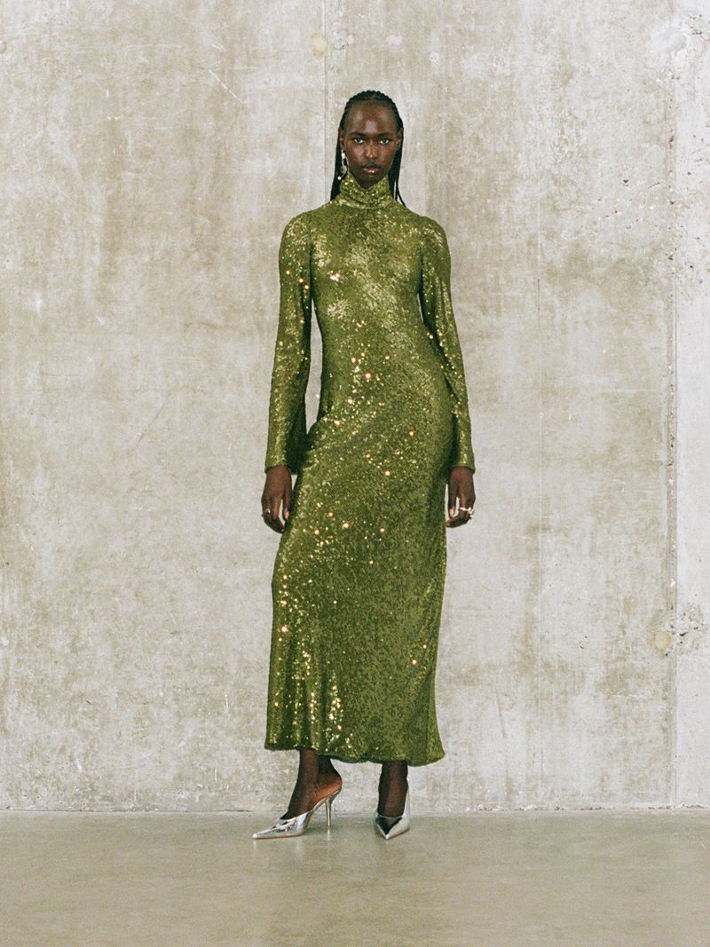 Elegant Black woman in shimmering green gown stands in front of a textured gray wall with stylish heels, creating a sophisticated fashion moment.