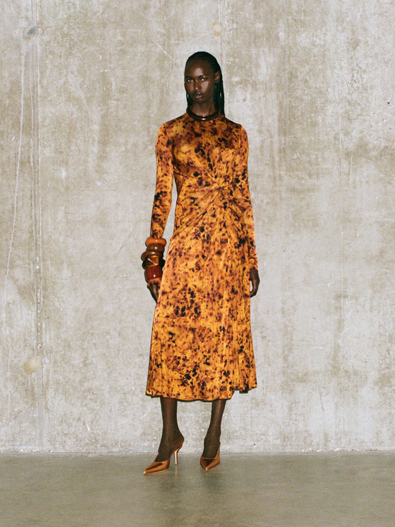 Adult Black female model wearing long-sleeve orange patterned dress, chunky bracelets, and heels against a textured wall backdrop.