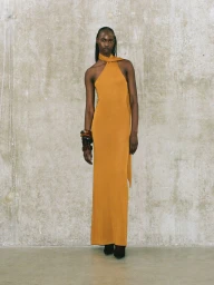 Model wearing a long yellow halterneck dress standing against a textured concrete wall.