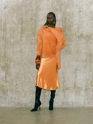 Model standing against a textured concrete wall wearing an orange textured sweater, satin midi skirt, and black boots.