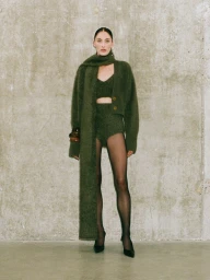 Model in olive green fuzzy knit set with matching long scarf standing against concrete wall texture