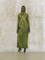 Model standing against a concrete wall wearing a long green sequined dress and metallic heeled shoes.