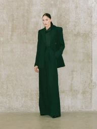 Model wearing a dark green structured blazer and matching wide-leg trousers against a weathered concrete wall background.