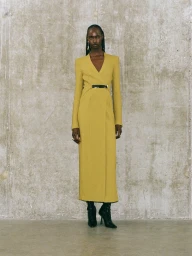 Model in a long yellow dress standing against a mottled grey concrete wall background.