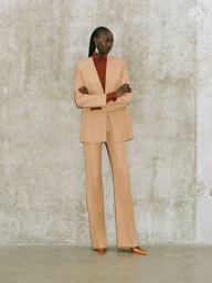 Model wearing a tan pantsuit and red top standing against a textured concrete wall for Galvan Pre-Spring 2025.