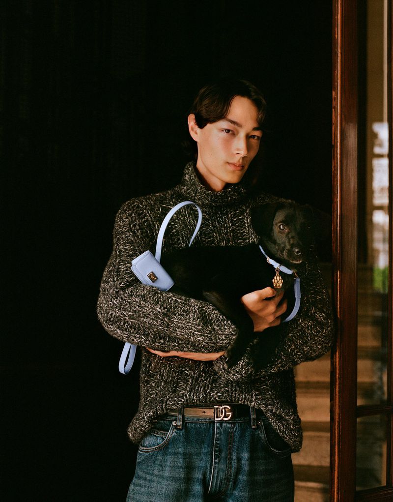 Adult male holding a black puppy with a light blue leash, a light blue pouch, wearing jeans and a gray sweater indoors.