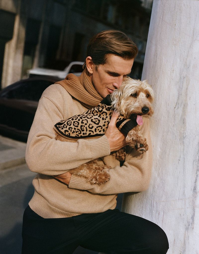 Man in tan sweater holds a small dog wearing a leopard-print coat outdoors on a sunny day, with cars and a building in the background.