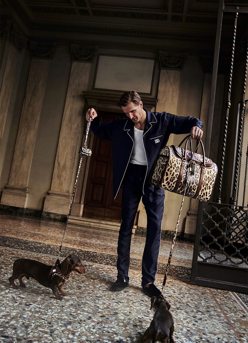 Man in pajamas poses with two dachshunds on leashes and a leopard print duffel bag inside an elegant, tiled hall.