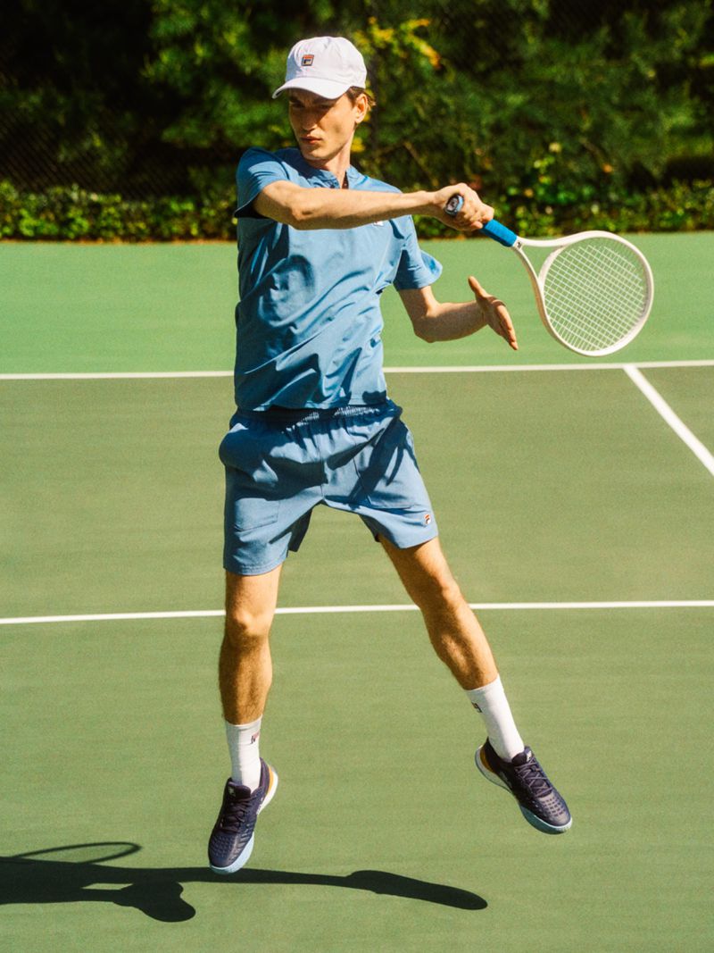 Man in motion playing tennis on outdoor court. He is wearing a blue athletic outfit, white cap, and navy tennis shoes.