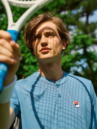 Man in blue shirt with tennis racket outdoors, sunlight casting shadow on face. Fila logo visible. Sports, athleticism, summer aesthetic.