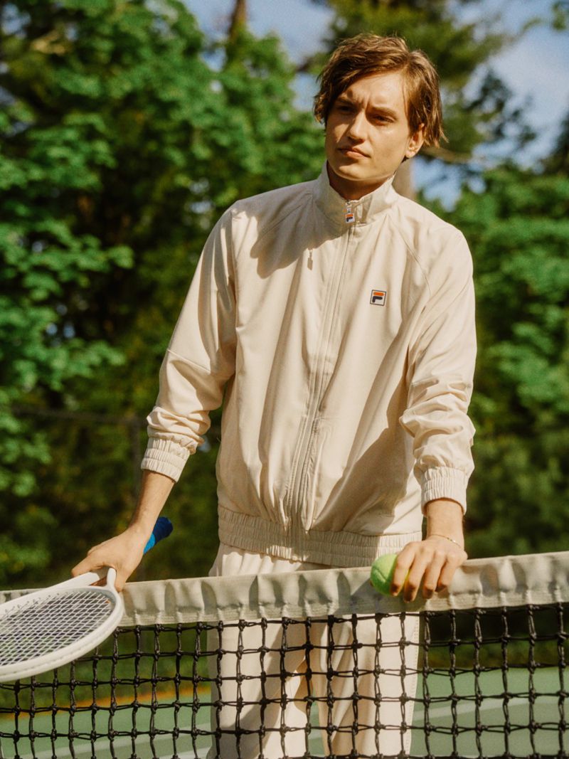Adult male in tennis outfit stands by the net on an outdoor court, holding a tennis ball and racket, with green trees in the background.