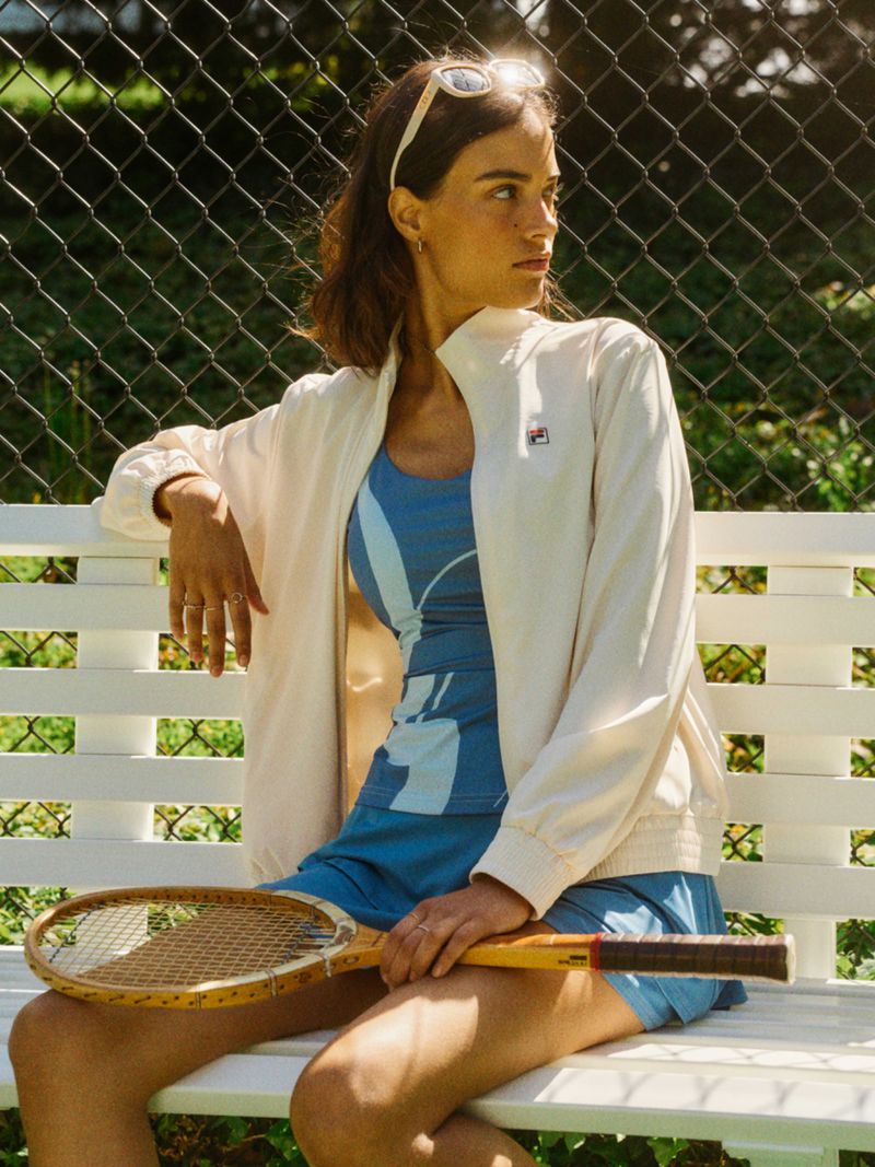 Adult woman in a tennis outfit with a racket sitting on a bench in front of a tennis court with a wire fence and green foliage.