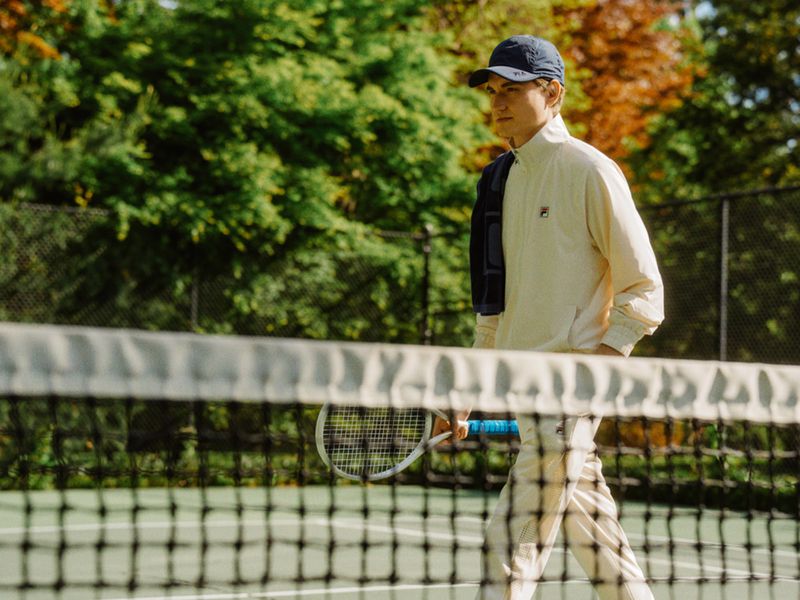 A young man wearing a cap and athletic wear walks across a green tennis court with net, trees in the background in outdoor setting.