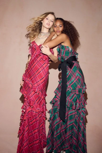 Two women in ruffled plaid gowns embrace against a soft pink backdrop in a fashion editorial, showcasing complementary color schemes and textures.