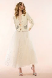 Model wearing a cream cardigan with sequin heart pockets and a tiered tulle skirt against a soft neutral studio backdrop.