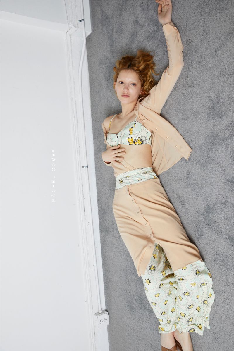 Red-haired young woman in a light peach colored outfit lying on a grey carpet and against a white wall in eye-level shot.