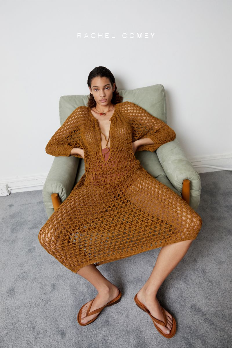Fashion shot of a woman in a crocheted dress sitting in a chair, posing in a studio with a minimal setup and wearing flip flops.