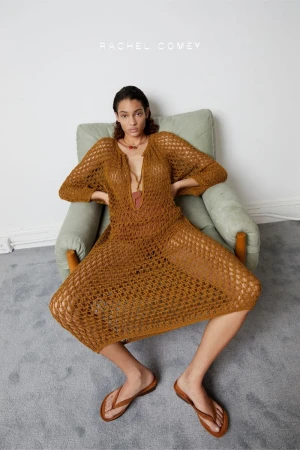 Fashion shot of a woman in a crocheted dress sitting in a chair, posing in a studio with a minimal setup and wearing flip flops.