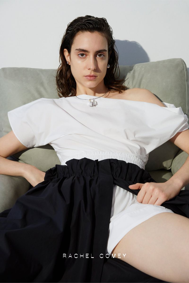 A stylish woman is posing indoors wearing a white blouse and a black skirt, seated on a green couch. The background is white.