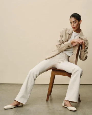 A woman in a tan jacket and off-white jeans sits on a wooden chair in a studio, her legs stretched out and arms resting behind her.
