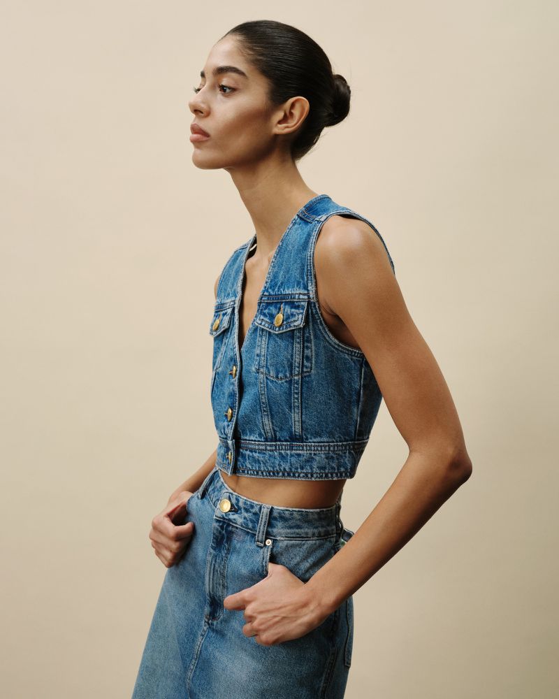 A woman in a denim vest and skirt poses in a studio with hands in pockets, showcasing a modern denim fashion ensemble.